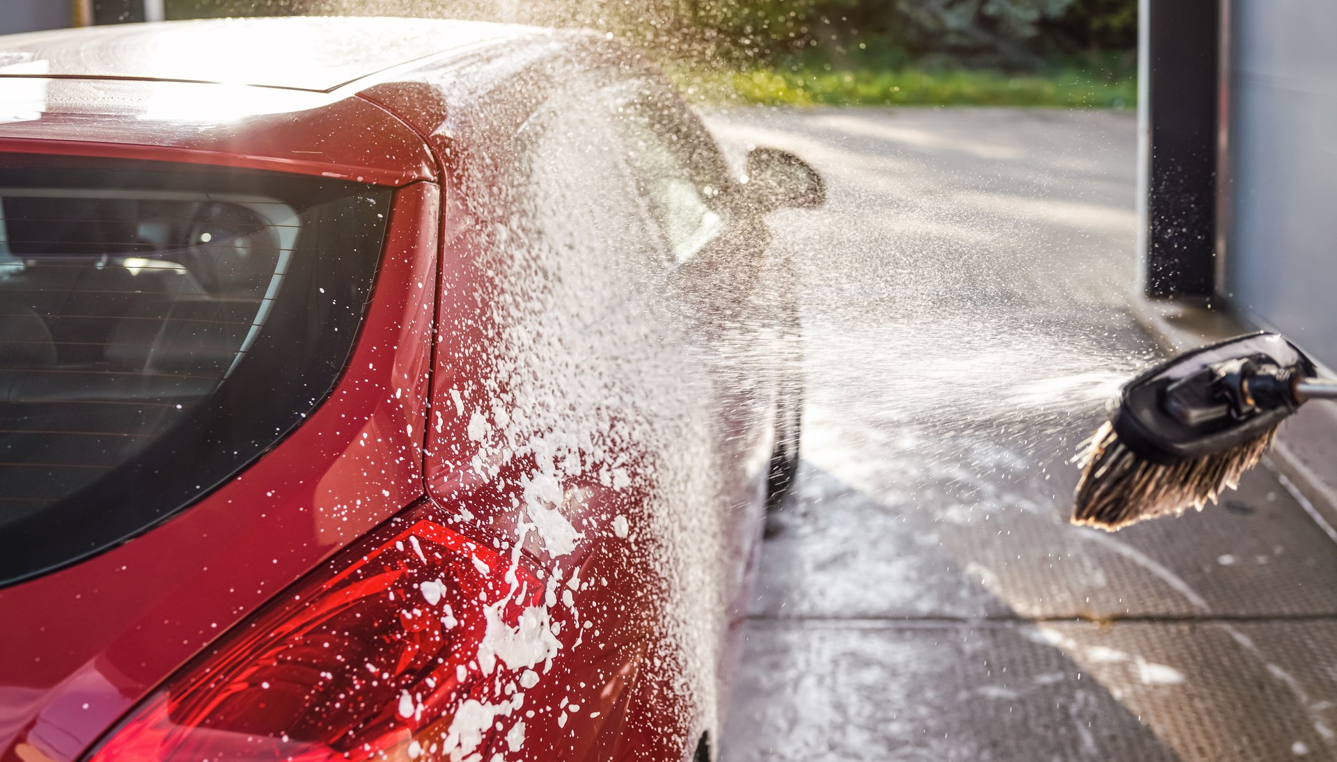 Red car washed in self serve carwash, detail on white soap foam spraying from brush broom onto side above rear wheel